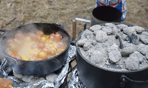 Two dutch ovens on a grate. One is open and shows a finished pot of potatoes, onions and cheese. The other pot is closed and has briquettes on top.