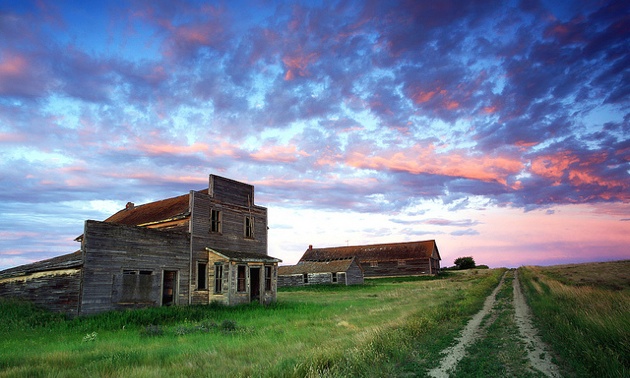 A Ghost Town in Saskatchewan with buildings on the left,  an old dirt tract on the right and a sky with clouds in different shades of pinks and reds.