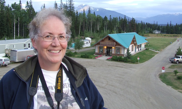 Pat wears a windbreaker and a camera on a blustery day. Behind her in the distance is the RV park's office.
