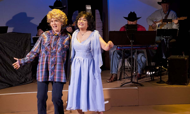 Two women are performing on a stage: one in a blue dress, the other in a plaid shirt and pants