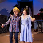 Two women are performing on a stage: one in a blue dress, the other in a plaid shirt and pants