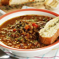 A bowl of hearty looking lentil stew is accompanied by a slice of french loaf and a spring of rosemary.