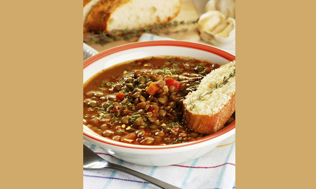 A bowl of hearty looking lentil stew is accompanied by a slice of french loaf and a spring of rosemary.