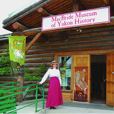 A woman dressed in a long red skirt and white blouse and hat stands in front of the MacBride Museum of Yukon History, ready to welcome visitors.
