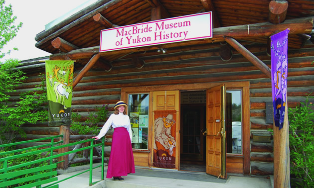 A woman dressed in a long red skirt and white blouse and hat stands in front of the MacBride Museum of Yukon History, ready to welcome visitors.