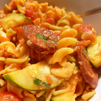 Saucy pasta is attractively arranged in a white bowl with pieces of chorizo, zucchini, and herbs.