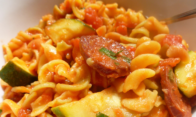 Saucy pasta is attractively arranged in a white bowl with pieces of chorizo, zucchini, and herbs.