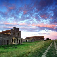 A Ghost Town in Saskatchewan with buildings on the left,  an old dirt tract on the right and a sky with clouds in different shades of pinks and reds.