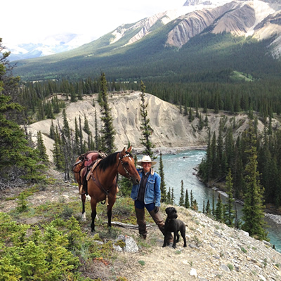 A woman standing above the river with her black dog and brown horse. 