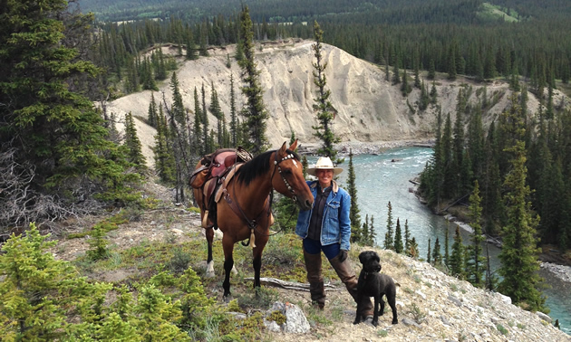 A woman standing above the river with her black dog and brown horse.