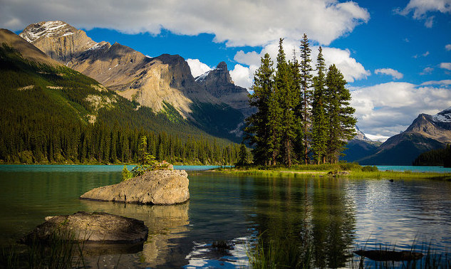 Spirit island in Maligne Lake is basking in the sun.  The waters from the island to the shore are calm and reflecting the island and mountains behind. 