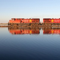 A CP train travelling across the rails with its reflection showing in a lake in the foreground. 