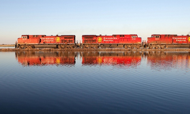 A CP train travelling across the rails with its reflection showing in a lake in the foreground. 
