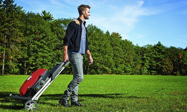 A young man walking through a park pulling a red TravelQ grill with him.