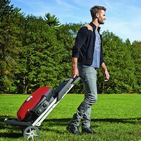 A young man walking through a park pulling a red TravelQ grill with him.